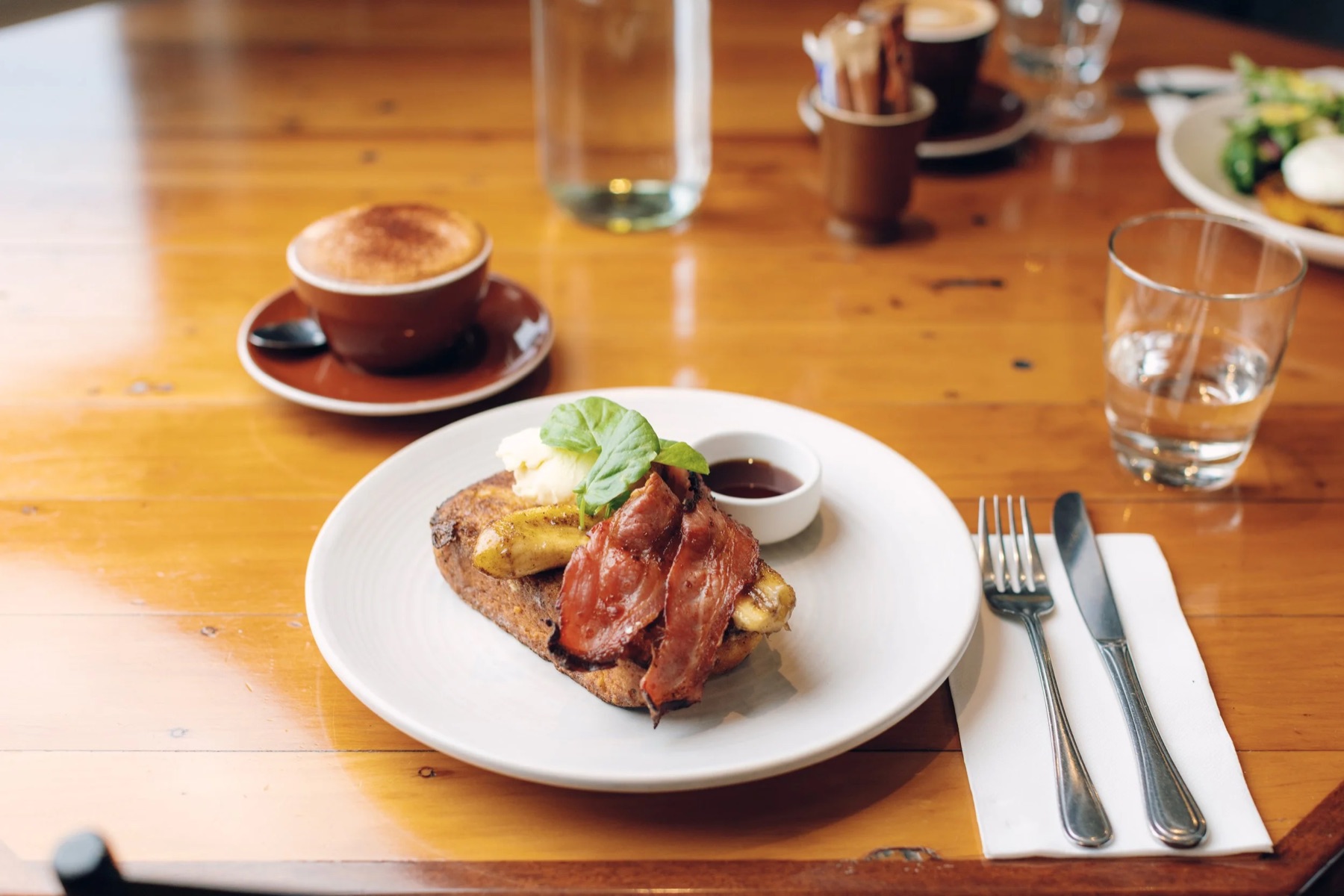 French toast with caramelised banana and streaky bacon beside a flat white on a honey-coloured timber table