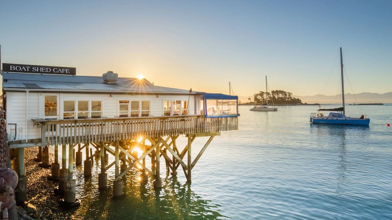 The white Boat Shed Cafe on its timber stilts over the water in Nelson at sunrise, yachts moored beyond