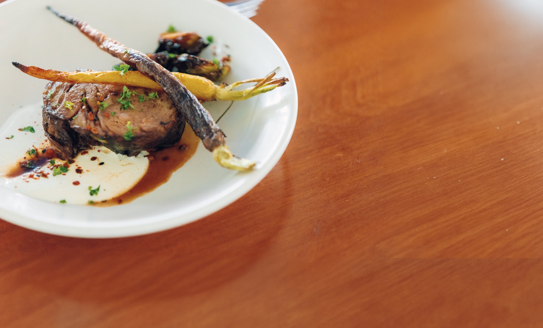 Braised lamb with heirloom carrots on a white plate, timber table beyond