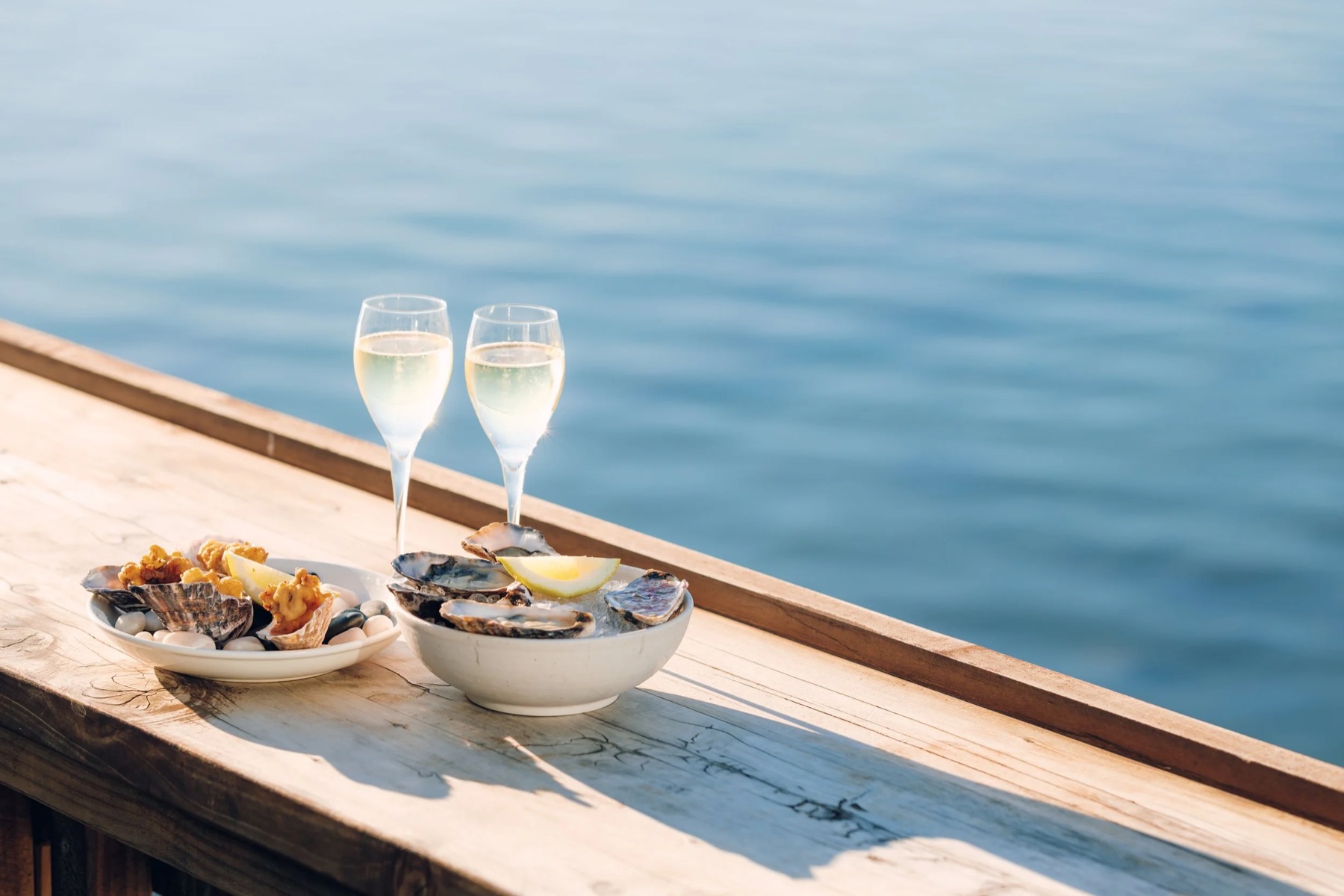 Two glasses of sparkling wine and a bowl of oysters on the deck rail in low sun, flat blue water behind